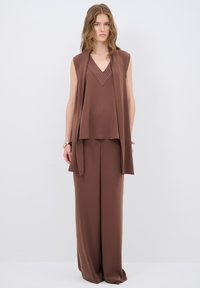 Bruine mouwloze top met een V-hals en decoratieve stitching, gecombineerd met losse bruine broek, beide gemaakt van glad stof.