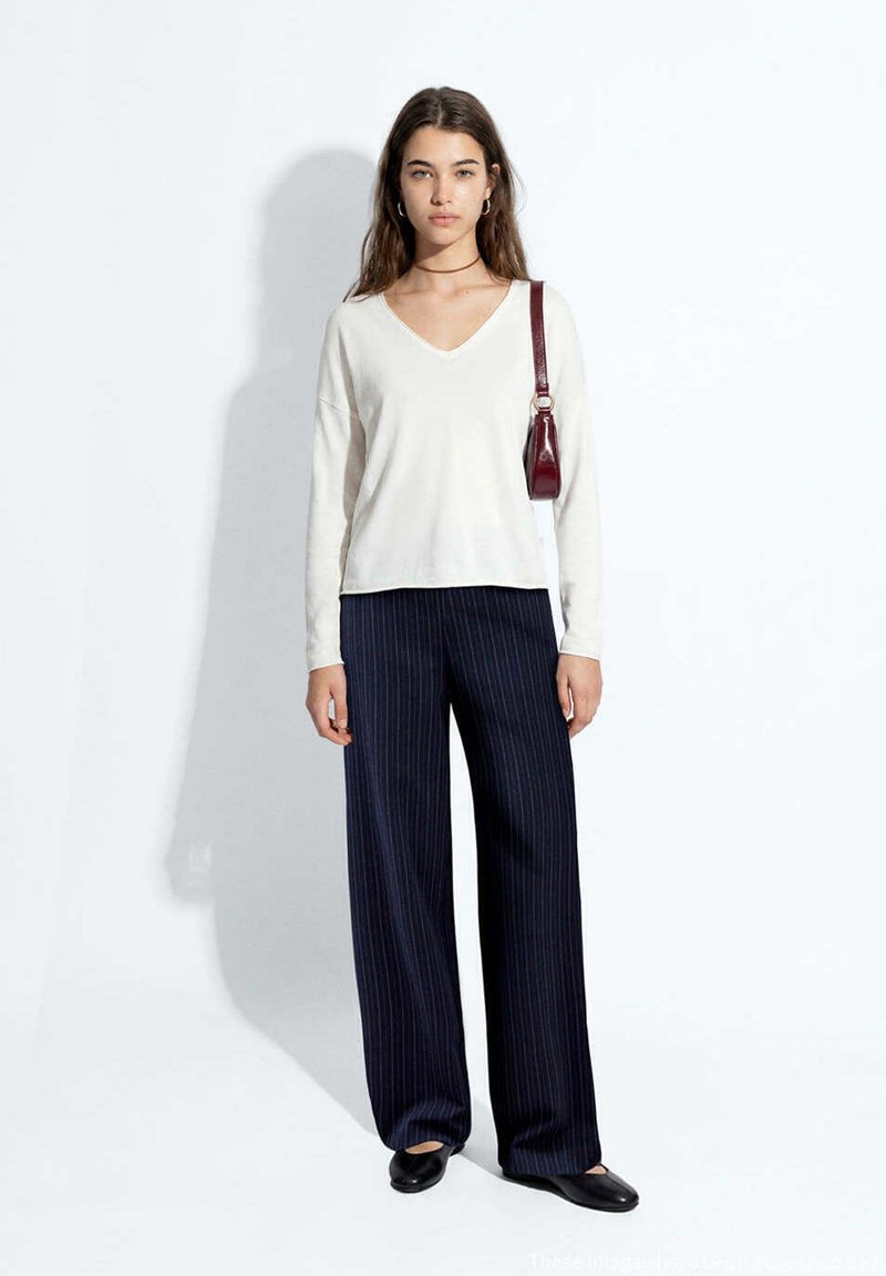 Femme portant un pull blanc à col en V, un pantalon large à fines rayures foncées, des ballerines noires et un petit sac à bandoulière bordeaux sur un fond uni.