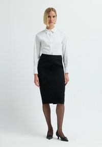 Weißes, kragenloses Hemd mit Knopfverschluss, kombiniert mit einem figurbetonten schwarzen Bleistiftrock, durchsichtigen Strumpfhosen und schwarzen spitzen High Heels.