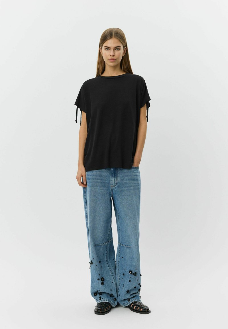 Femme debout portant un haut ample noir, un jean bleu à jambes larges avec des ornements floraux noirs près des poignets, et des chaussures tissées noires sur fond blanc.