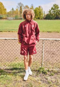 Bordeaux- og gule aksenter med Arsenal-logo; inkluderer matchende shorts og hvite sneakers. Grassterreng og gjerde i bakgrunnen.