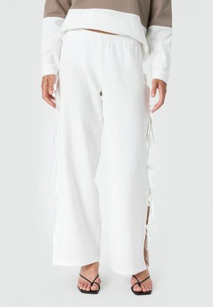 Pantalon blanc à jambes larges avec liens latéraux porté par une personne en sandales noires à brides et un haut beige et blanc à manches longues.