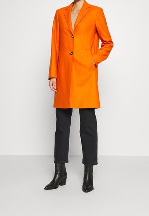 Persoon draagt een feloranje knielange jas over een zwarte broek en zwarte laarzen met hakken, staat met één hand in de zak tegen een witte achtergrond.