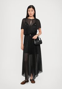 Zwarte doorzichtige maxi-jurk met korte mouwen, voorzien van een keyhole-halslijn, subtiele glans en een ceintuur in de taille. Gecombineerd met slippers in luipaardprint.