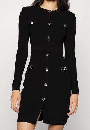 Schwarzes enganliegendes Langarmkleid mit silbernen Knöpfen vorne, zwei Brusttaschen mit Knöpfen und zwei Taillenschnallen, getragen von einer Frau.