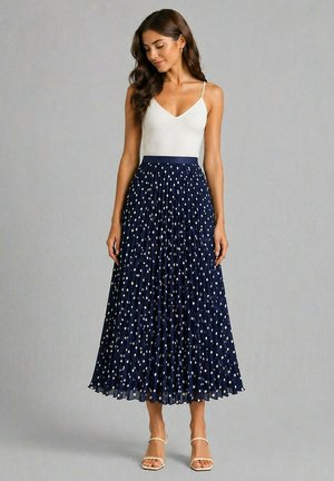 Femme portant un haut blanc sans manches et une jupe midi plissée bleu marine à pois blancs, debout devant un fond gris uni.