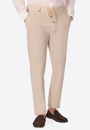 Uomo che indossa pantaloni beige con coulisse, camicia bianca a maniche lunghe e scarpe slip-on marrone scuro, mani in tasca.