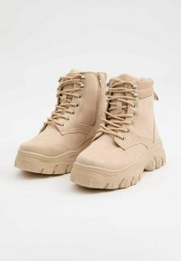 Botas de ante beige hasta el tobillo con suelas gruesas y dentadas, cordones en la parte delantera, cremalleras laterales y cuellos acolchados sobre un fondo blanco.