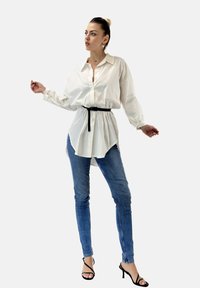 Camicia bianca a maniche lunghe con una vestibilità morbida, cintrata in vita con una cintura nera, abbinata a jeans skinny azzurri e tacchi a strappi neri.