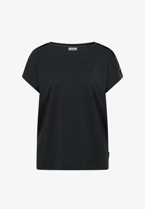 Personne souriante portant un t-shirt noir à manches courtes avec un détail de sangle à boucle argentée sur la manche et un chapeau seau en jean bleu clair.