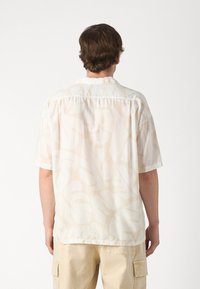 Uomo che indossa una camicia beige a maniche corte con stampa tropicale e pantaloni cargo beige, in piedi di spalle contro uno sfondo bianco semplice.