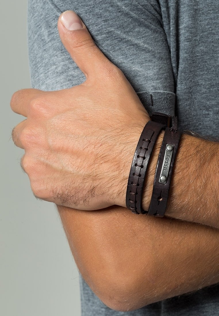 Bras d’homme croisé sur la poitrine portant une chemise grise et un bracelet en cuir marron foncé Fossil au poignet.
