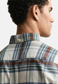 Camicia abbottonata con motivo a quadri in tonalità di blu, verde, marrone e crema. Tessuto in cotone, colletto corto con chiusura a bottone sul retro.