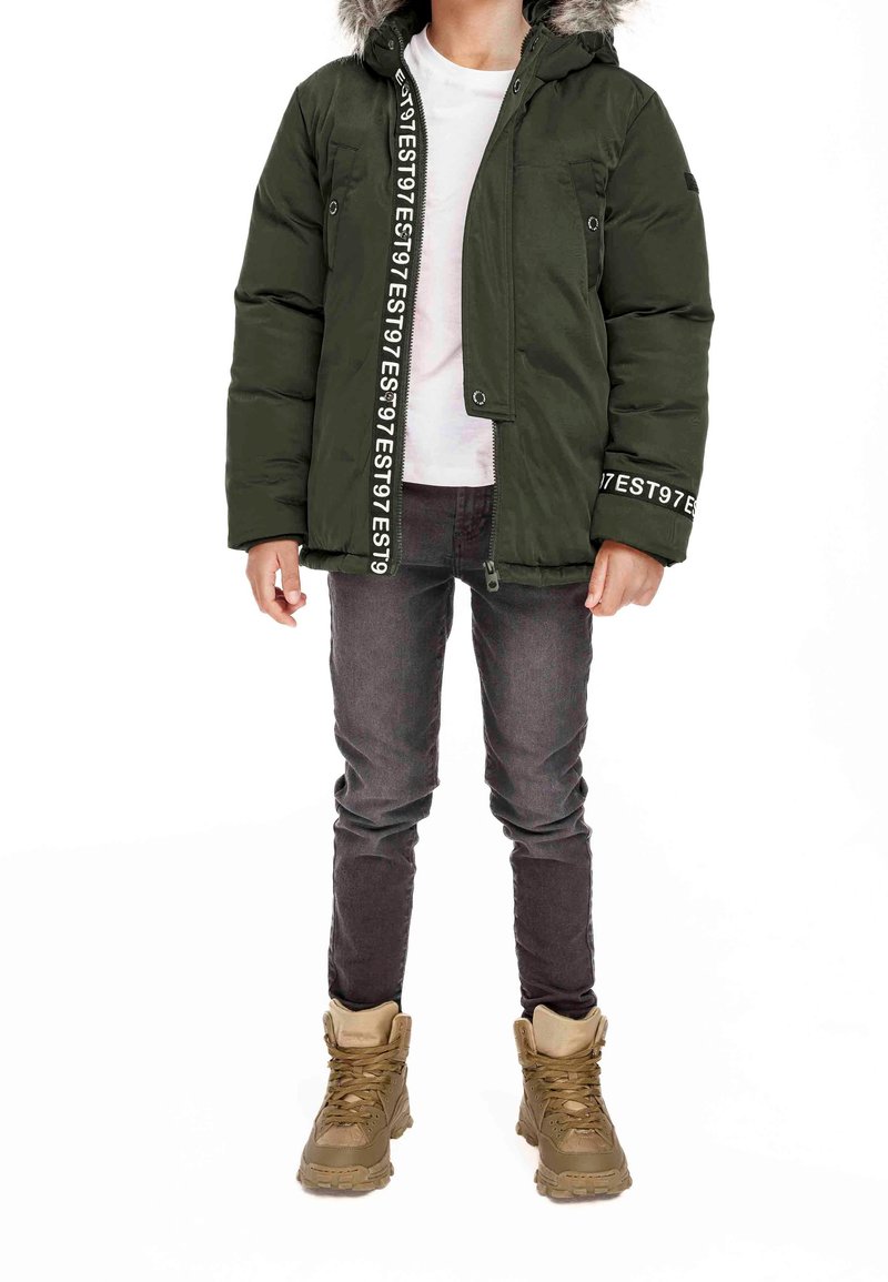 Enfant portant une veste d'hiver matelassée verte avec capuche en fourrure, pantalon noir et bottes beige, debout sur fond blanc.