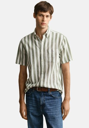 Junger Mann trägt ein grün-weiß vertikal gestreiftes kurzärmeliges Hemd, in blaue Jeans mit braunem Gürtel gesteckt, steht vor weißem Hintergrund.