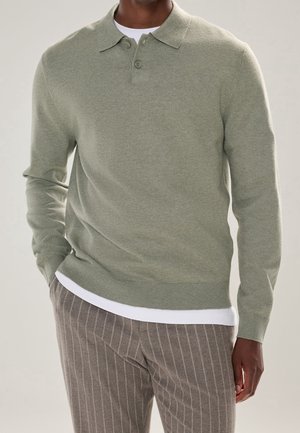 Uomo che indossa un maglione polo a maniche lunghe verde chiaro sopra una camicia bianca con pantaloni grigi a righe sottili, mano in tasca, su sfondo neutro.