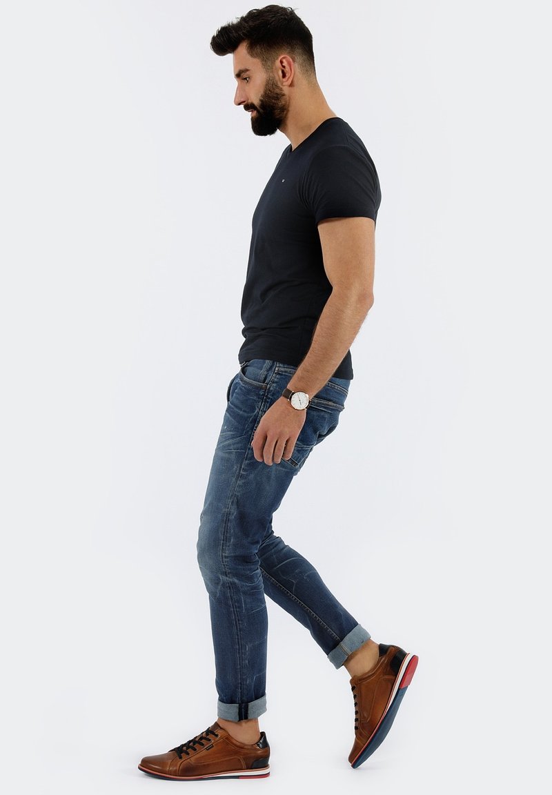 Dunkelgraues V-Ausschnitt-T-Shirt, blaue Jeans mit umgeschlagenen Säumen und braune Sneakers mit roten Akzenten. Das Modell posiert seitlich.