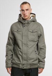 Chaqueta con capucha de color verde oliva, con forro de forro polar, dos bolsillos en el pecho y cierres de botones a presión. Presenta puños y dobladillo acanalados.