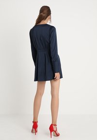 Vestido azul marino con mangas largas, cintura entallada y falda plisada. Combinado con zapatos de tacón rojo, que exhiben una textura elegante y suave.