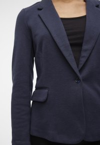 Blazer bleu marine en tissu texturé avec une fermeture à un bouton, des revers crantés et une poche poitrine. Porté sur un haut noir.