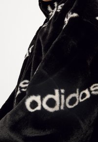 Černá fleecová bunda s kapucí, charakterizovaná měkkou texturou, volným střihem a bílým logem "adidas" výrazně umístěným na rukávu.