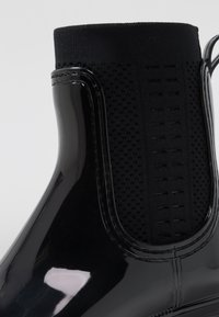 Black glossy ankle boot with elastic mesh sides, rubber sole, and textured accents. Smooth finish with a sleek, modern silhouette.