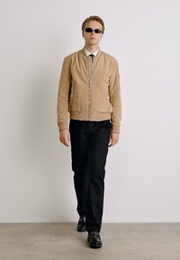 Veste aviateur beige avec une fermeture éclair à l'avant, deux poches latérales, pantalon noir ajusté et chaussures noires, portée avec une chemise blanche et une cravate.