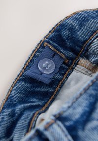 Verstelbare blauwe elastische tailleband met doorzichtige plastic knoop op blauwe spijkerbroek, met bruine stiksels en witte binnenvoering.