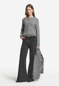 Grå stickad cardigan med rund hals och knäppning, i kombination med högmidjade svarta jeans. Modellen håller en grå vadderad jacka.