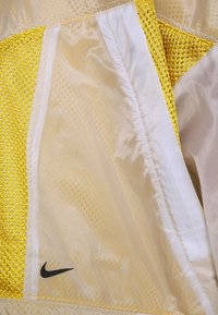 Gul nätjacka med glänsande vita paneler och en svart Nike-logga. Lättviktsmaterial, strukturerad in- och utsida samt synlig sömnad.