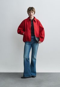 Άτομο που φοράει κόκκινο μπουφάν με φερμουάρ, μαύρο πουκάμισο, μπλε τζιν καμπάνα και μαύρα παπούτσια, κρατώντας μια μικρή μαύρη τσάντα.