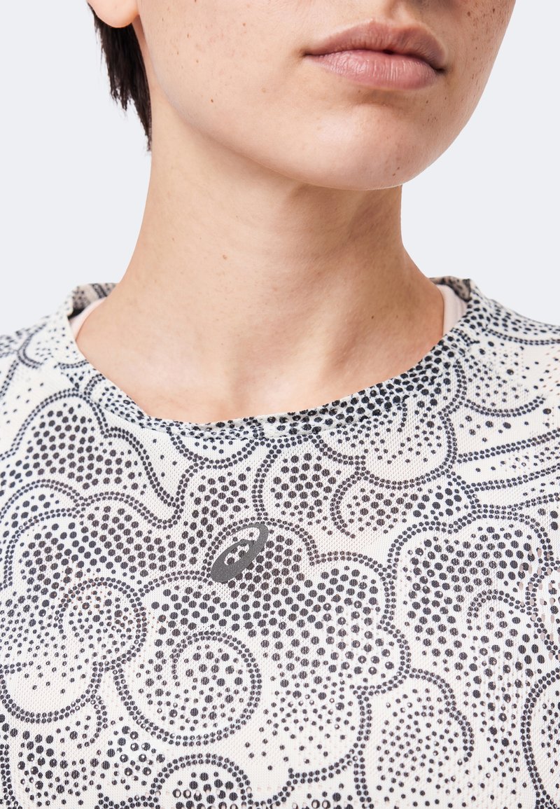 Nahaufnahme des unteren Gesichts und des Halses einer Person, die ein weißes Oberteil mit kunstvollen schwarzen gepunkteten Blumenmustern und einem kleinen Logo trägt.