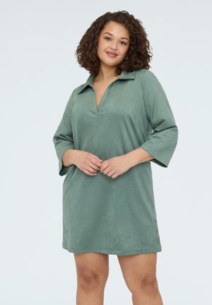 Vrouw met krullend haar, gekleed in een los, lichtgroen jurk met driekwart mouwen en een kraag, staand tegen een effen witte achtergrond.