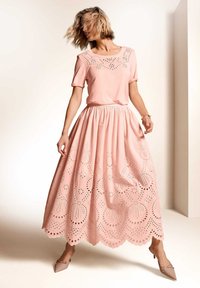 Ein hellrosa Kleid mit einem figurbetonten Oberteil und einem ausgestellten Rock, der mit Lochstickerei verziert ist. Der Rock hat einen gewellten Saum und eine dezente Textur.