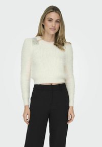 White, fluffy cropped sweater with long sleeves, featuring silver embellishments on the shoulders, paired with black high-waisted trousers.
