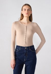 Haut beige à manches longues avec un décolleté en forme de scoop et une patte de boutons à l'avant. Associé à un jean foncé taille haute, avec des coutures contrastées.