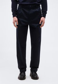 Άνδρας που φοράει μαύρο παντελόνι ευθείας γραμμής, μαύρα μοκασίνια και μαύρο μπλουζάκι μακρυμάνικο που είναι μπηγμένο μέσα, στέκεται μπροστά σε ένα απλό φόντο.