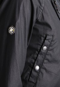 Veste légère noire avec une finition texturée, présentant un patch logo hexagonal sur la manche et des boutons-pression double boutonnage sur le côté.