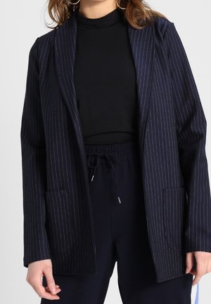 Femme portant un blazer bleu marine à fines rayures sur un col roulé noir, accompagné d'un pantalon bleu marine avec cordon de serrage.