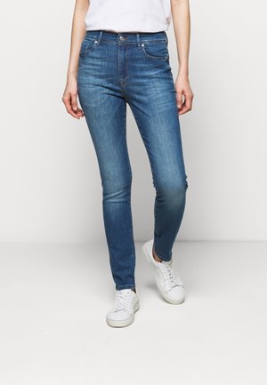 Person iført blå slim-fit jeans og hvide sneakers, stående mod en ensfarvet hvid baggrund.