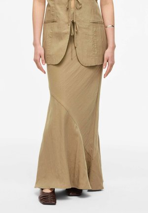 Femme portant une jupe beige avec une couture diagonale, un gilet assorti noué, un bracelet en argent et des chaussures marron froncées à bout carré sur fond blanc.