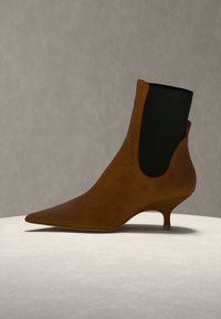 Bottine en daim marron à bout pointu, panneau latéral élastique noir et talon bas incurvé sur une surface claire sur fond neutre.