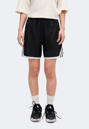 Person iført sorte sportsshorts med hvid kant, hvide sokker, sorte atletiksk sko og en løs hvid kortærmet skjorte, der står oprejst.