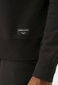 Sudadera negra con textura acanalada y un parche con el logo que dice "BJÖRN BORG STOCKHOLM 1956" cerca del dobladillo. Diseño sencillo y moderno.