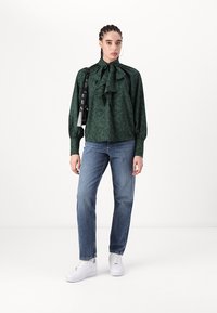 Blusa verde a fantasia con dettaglio fiocco, abbinata a jeans dritti blu medio e sneaker bianche. Borsa nera come accessorio.