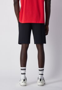 Pantalones cortos negros con un corte relajado, combinados con calcetines blancos con rayas negras y zapatillas. Camiseta roja no completamente visible. Fondo liso.
