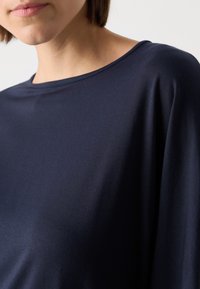 Top blu navy con scollatura ampia, texture del tessuto liscia e vestibilità morbida. Ingrandimento del tessuto che scende su una spalla.