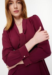 Burgundská dlhá sukňa s viazaním pri krku, štruktúrovaná tkanina a gombíkové akcenty na rukávoch. Model s prekríženými rukami, neutrálny pozadie.