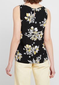 Top senza maniche nero con motivo floreale in bianco e giallo, caratterizzato da un dettaglio a chiave sulla schiena e una texture leggera e liscia.