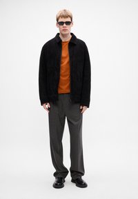 Giovane uomo che indossa occhiali da sole neri, giacca nera, maglietta arancione, pantaloni grigi a quadri e scarpe nere su uno sfondo bianco uniforme.